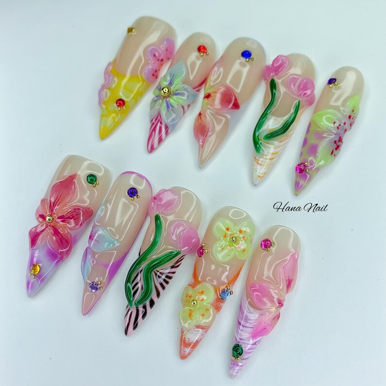 May include: A set of ten artificial nails with a floral design. Each nail is a different shape and features a clear base with 3D flowers in various colors, including pink, yellow, and green. Some nails have small gems. The text "Hana Nail" is visible.