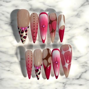 May include: A set of ten long, pointed false nails. Designs include leopard print, pink gradients, polka dots, and a starburst design with a pink gem. Some nails feature brown and white patterns. The nails are arranged on a white marble surface.