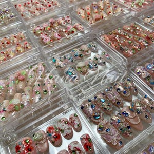 May include: A selection of acrylic nails displayed in clear plastic cases. The nails showcase diverse designs, such as floral patterns, rhinestones, and butterfly motifs. Colours include pink, white, red, and iridescent gems. The nails are arranged in rows.