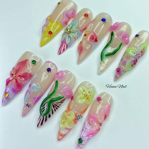 May include: A set of ten long, almond-shaped artificial nails. Each nail features a nude base with 3D floral designs in various colors, including pink, yellow, and green. Some nails have small gem accents. The text "Hana Nail" is visible.