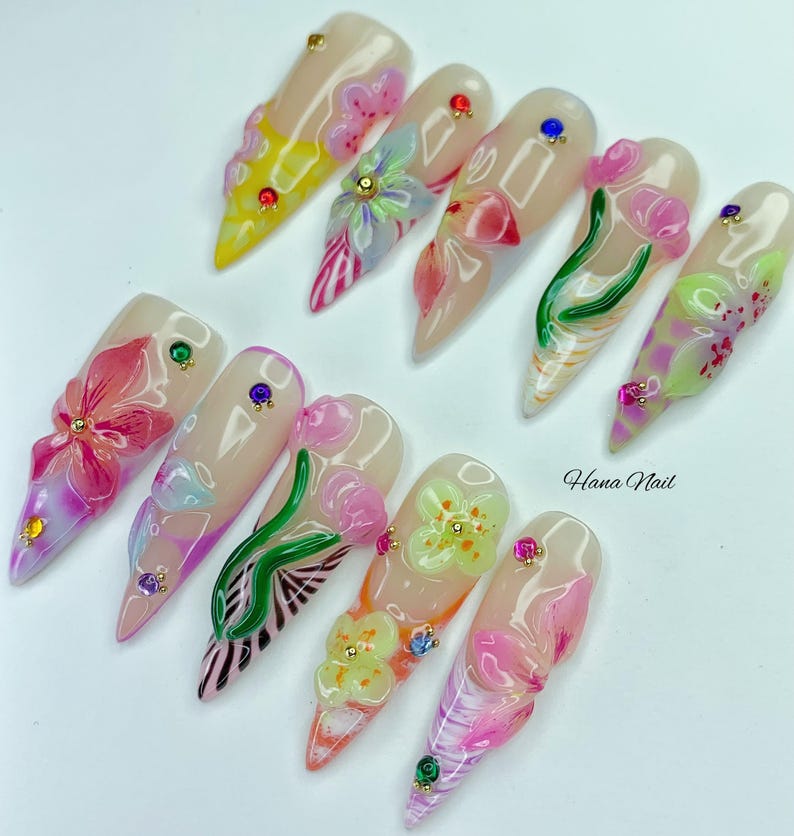 May include: A set of ten long, almond-shaped artificial nails. Each nail features a nude base with 3D floral designs in various colors, including pink, yellow, and green. Some nails have small gem accents. The text "Hana Nail" is visible.