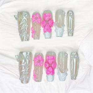 Puede incluir: Un juego de diez u&ntilde;as artificiales con acabado nacarado. Algunas u&ntilde;as presentan dise&ntilde;os de mariposas y flores en 3D en rosa y plata. Otras u&ntilde;as tienen un color gris claro liso con detalles de perlas.