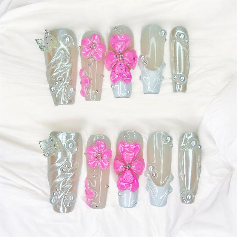 Puede incluir: Un juego de diez u&ntilde;as artificiales con acabado nacarado. Algunas u&ntilde;as presentan dise&ntilde;os de mariposas y flores en 3D en rosa y plata. Otras u&ntilde;as tienen un color gris claro liso con detalles de perlas.