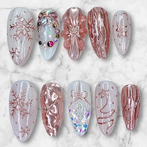May include: A collection of ten artificial nails with various designs. Some nails feature rose gold snowflakes, a flower, and a Christmas tree. Others have textured patterns and sparkling embellishments. The nails are oval-shaped and set against a marble background.