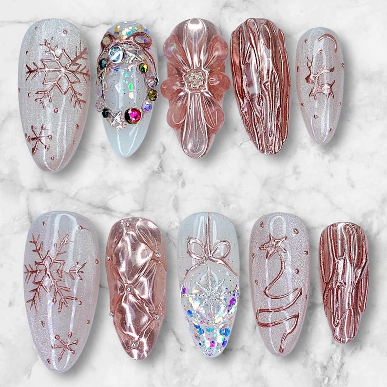 May include: A collection of ten artificial nails with various designs. Some nails feature rose gold snowflakes, a flower, and a Christmas tree. Others have textured patterns and sparkling embellishments. The nails are oval-shaped and set against a marble background.