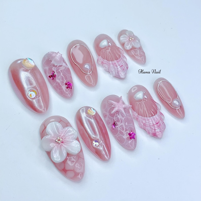 Peut inclure: Ensemble de dix ongles press&eacute;s en forme d'amande rose et blanc avec diff&eacute;rents motifs, notamment des coquillages, des fleurs et des paillettes. Les ongles sont d&eacute;cor&eacute;s de paillettes roses, blanches et iris&eacute;es. Le texte "Hana Nail" est visible sur l'image.