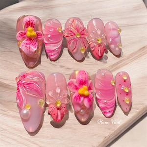 May include: Pink and white oval-shaped fake nails with 3D floral designs. Some nails have small pearl accents. The text "Hana Nail" is visible in the bottom right corner of the image.