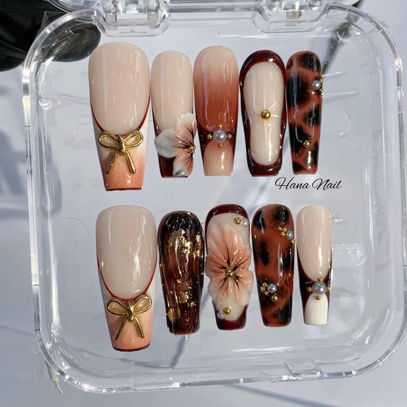 May include: A set of ten artificial nails in a clear plastic case. The nails feature various designs, including a bow, floral accents, and a leopard print pattern. Colors include nude, brown, gold, and white. The text "Hana Nail" is visible on the case.