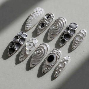 May include: A collection of ten white and silver nail designs. The nails feature various patterns, including spirals, geometric shapes, and eye-like designs. The silver accents create a striking contrast against the white base, showcasing intricate details.
