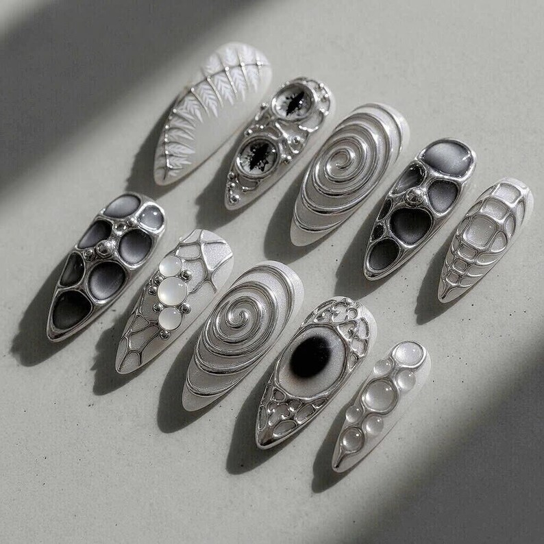 May include: A collection of ten white and silver nail designs. The nails feature various patterns, including spirals, geometric shapes, and eye-like designs. The silver accents create a striking contrast against the white base, showcasing intricate details.