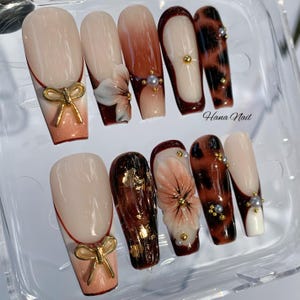 May include: A set of ten artificial nails in various designs. Some nails feature a nude base with a gold bow accent, while others showcase floral designs, leopard print, and gold leaf details. The nails are in a clear plastic container.