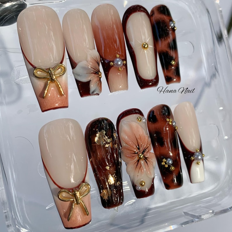 May include: A set of ten artificial nails in various designs. Some nails feature a nude base with a gold bow accent, while others showcase floral designs, leopard print, and gold leaf details. The nails are in a clear plastic container.