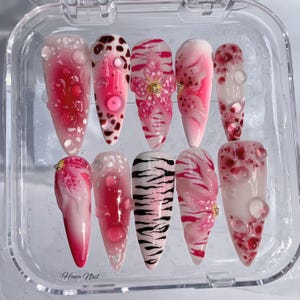 May include: A set of ten artificial nails in a clear plastic case. The nails feature various designs, including pink and white swirls, floral accents, and a zebra print. Some nails have 3D bubble-like embellishments and small gold accents. The text "Hana Nail" is visible.