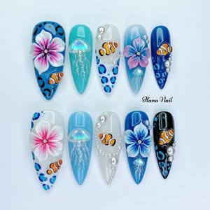 May include: A collection of ten long, almond-shaped artificial nails with marine-inspired designs. Each nail showcases a unique design, including flowers, jellyfish, clownfish, and pearl embellishments. The nails are painted in shades of blue, white, and black.