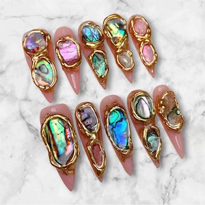 May include: A set of ten stiletto-shaped press-on nails with a light pink base. Each nail features irregular, iridescent abalone shell fragments encased in raised, textured gold metallic borders that follow the organic shapes of the shells.