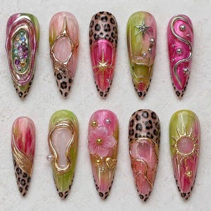 May include: A set of ten almond-shaped press-on nails featuring a mix of pink and lime green marbled bases, leopard print tips, and 3D metallic gold and silver accents including stars, flowers, hearts, and small rhinestones in various colors.