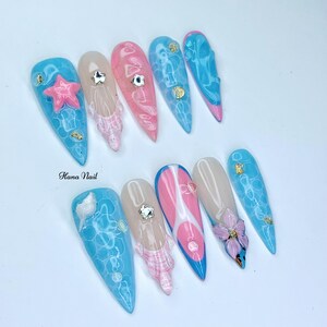 Puede incluir: Un conjunto de diez u&ntilde;as postizas largas y puntiagudas con una variedad de dise&ntilde;os. Las u&ntilde;as est&aacute;n pintadas en tonos de azul, rosa y blanco. Algunas u&ntilde;as tienen un dise&ntilde;o 3D, como una estrella de mar, una flor o una concha. Las u&ntilde;as est&aacute;n dispuestas en dos filas sobre una superficie blanca. Hana Nail est&aacute; escrito en texto negro sobre la superficie.