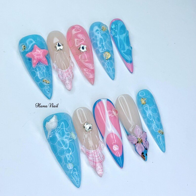 Puede incluir: Un conjunto de diez u&ntilde;as postizas largas y puntiagudas con una variedad de dise&ntilde;os. Las u&ntilde;as est&aacute;n pintadas en tonos de azul, rosa y blanco. Algunas u&ntilde;as tienen un dise&ntilde;o 3D, como una estrella de mar, una flor o una concha. Las u&ntilde;as est&aacute;n dispuestas en dos filas sobre una superficie blanca. Hana Nail est&aacute; escrito en texto negro sobre la superficie.