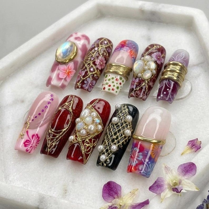 May include: A set of ten press-on nails featuring diverse designs including floral patterns, gold filigree, pearl embellishments, iridescent cabochons, and metallic bands. The nails display a variety of colours such as pink, deep red, black, and purple.