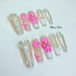 Puede incluir: Conjunto de diez u&ntilde;as artificiales largas en forma de ata&uacute;d. Las u&ntilde;as presentan una base nacarada con detalles florales y lazos rosas, dijes de mariposa plateados y peque&ntilde;as perlas. El texto "Hana Nail" es visible.
