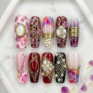 May include: A set of ten press-on nails featuring diverse designs including pink and red floral patterns, gold wire filigree, iridescent cabochons, white pearls, and metallic bands. The nails display a mix of glossy finishes and intricate textures.