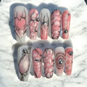 May include: A set of ten long, coffin-shaped artificial nails. The nails feature a variety of designs, including pink and white gradients, silver metallic embellishments, and textured patterns. Some nails have a heart shape, while others have a swirl or bubble design.
