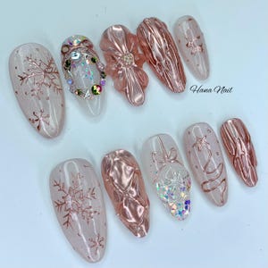 May include: A collection of artificial nails with a winter theme. The nails feature a variety of designs, including snowflakes, textured surfaces, and decorative embellishments. Colors include pink, clear, and rose gold. The text "Hana Nail" is visible.