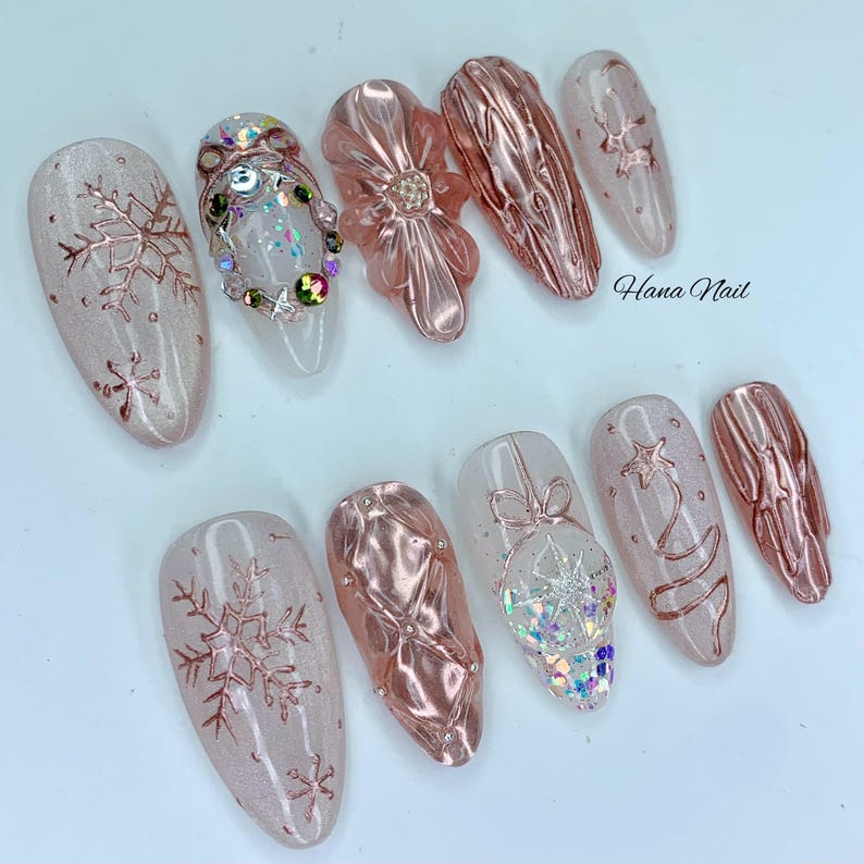 May include: A collection of artificial nails with a winter theme. The nails feature a variety of designs, including snowflakes, textured surfaces, and decorative embellishments. Colors include pink, clear, and rose gold. The text "Hana Nail" is visible.