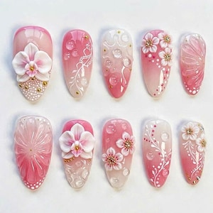 May include: A collection of ten artificial nails showcasing diverse designs. The nails display a pink and white gradient, embellished with 3D flowers, gold details, and delicate patterns. Some nails have a textured, bubble-like effect.