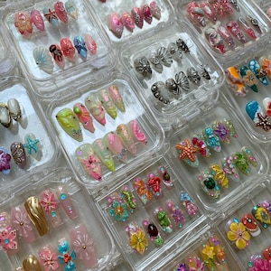Könnte beinhalten: Eine Sammlung verschiedener Sets mit aufklebbaren Nägeln in transparenten Kunststoffbehältern. Die Nägel weisen verschiedene Designs auf, darunter Blumen-, Muschel- und abstrakte Muster, mit Farben wie Rosa, Weiß und Gold. Einige Nägel haben 3D-Verzierungen.