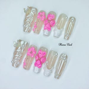 Puede incluir: Un conjunto de u&ntilde;as artificiales con diversos dise&ntilde;os. Algunas u&ntilde;as est&aacute;n adornadas con adornos florales rosas en 3D, mientras que otras presentan detalles de perlas y mariposas. El texto "Hana Nail" es visible.