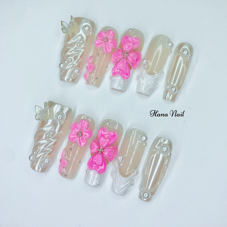Puede incluir: Un conjunto de u&ntilde;as artificiales con diversos dise&ntilde;os. Algunas u&ntilde;as est&aacute;n adornadas con adornos florales rosas en 3D, mientras que otras presentan detalles de perlas y mariposas. El texto "Hana Nail" es visible.
