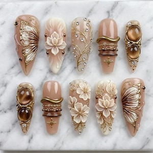 May include: A collection of ten almond-shaped artificial nails. The nails feature various designs, including floral patterns, butterfly motifs, and gold embellishments with rhinestones. The color palette includes shades of beige, brown, and gold.