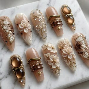 May include: A collection of ten almond-shaped artificial nails. The nails feature various designs, including floral embellishments, gold accents, and jewel-like adornments. Colors include nude, gold, brown, and white. Some nails have a butterfly wing design.