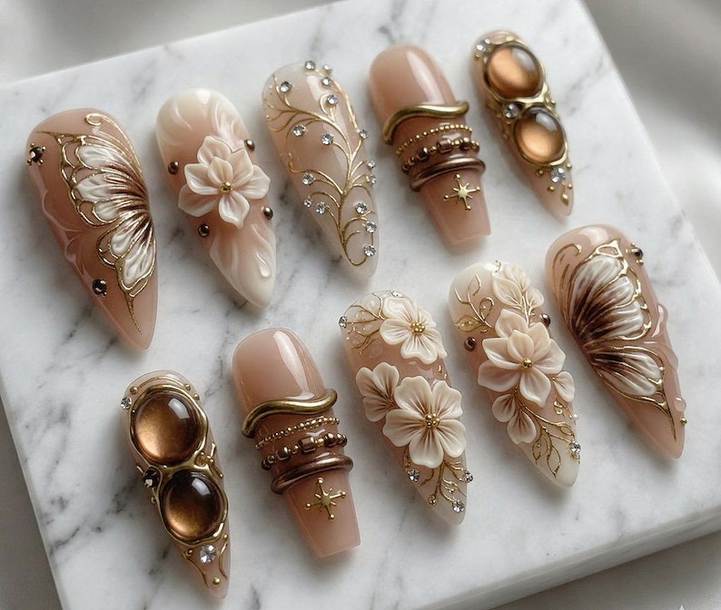 May include: A collection of ten almond-shaped artificial nails. The nails feature various designs, including floral embellishments, gold accents, and jewel-like adornments. Colors include nude, gold, brown, and white. Some nails have a butterfly wing design.