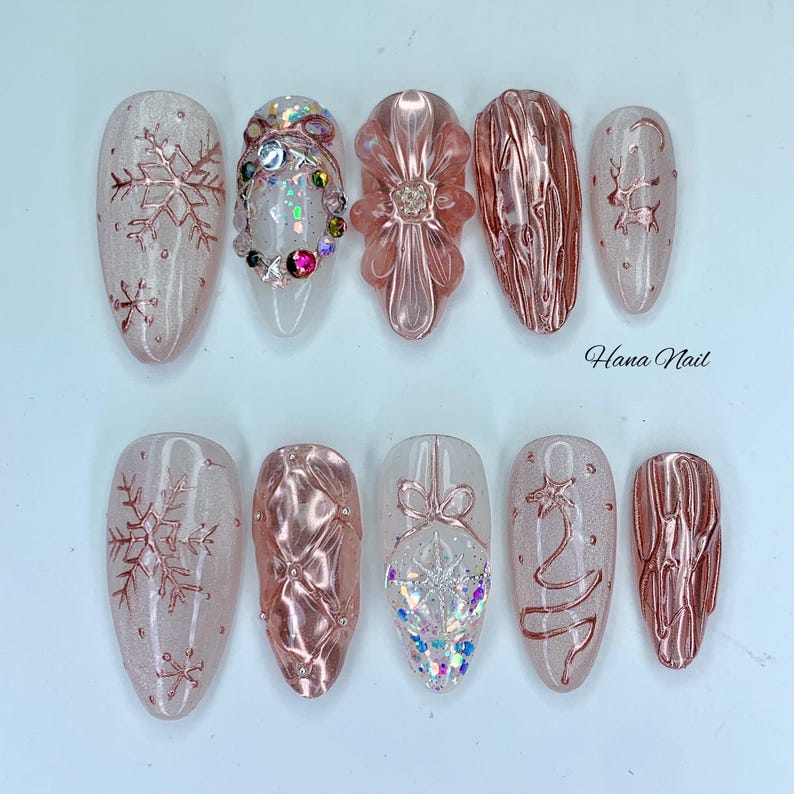 May include: A set of ten artificial nails with various designs. The nails feature a light pink base with rose gold accents, including snowflakes, floral patterns, and textured designs. Some nails are adorned with iridescent gems and glitter. The text "Hana Nail" is visible.