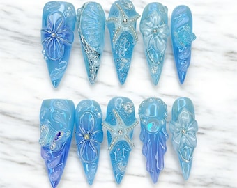 May include: A set of ten press-on nails in varying shades of blue, featuring 3D embellishments including sculpted flowers, starfish, silver beads, and iridescent accents. The almond-shaped nails display intricate textures and glossy finishes.