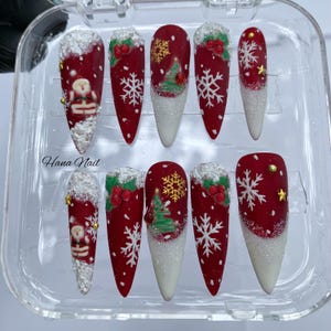 May include: Set of ten red and white Christmas-themed artificial nails. Designs include Santa Claus, snowflakes, Christmas trees, and gold stars. The nails are in a clear plastic container. The text "Hana Nail" is visible.