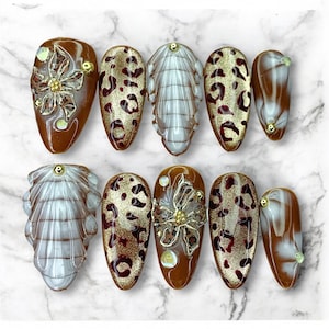 May include: A set of ten artificial nails in various designs. Some nails feature a brown base with gold floral accents, while others have a leopard print pattern. Additional nails have a white and brown swirl design. Each nail is oval-shaped.