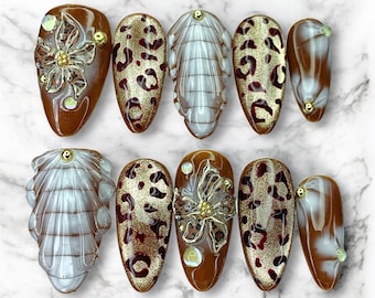 May include: A set of ten artificial nails in various designs. Some nails feature a brown base with gold floral accents, while others have a leopard print pattern. Additional nails have a white and brown swirl design. Each nail is oval-shaped.