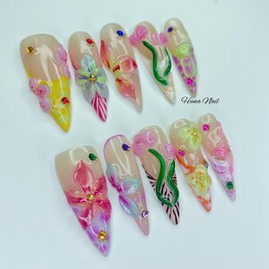 May include: A set of ten long, almond-shaped artificial nails. Each nail features a floral design with pink, yellow, and green accents, adorned with rhinestones. The text "Hana Nail" is visible on the right side.