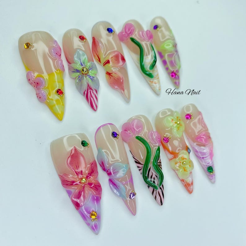 May include: A set of ten long, almond-shaped artificial nails. Each nail features a floral design with pink, yellow, and green accents, adorned with rhinestones. The text "Hana Nail" is visible on the right side.