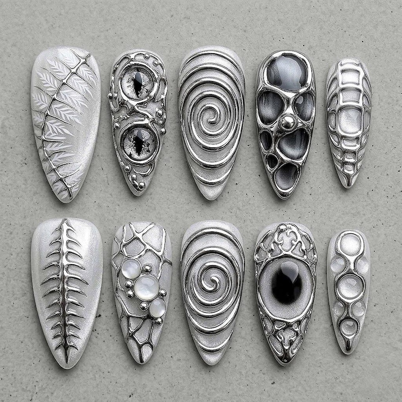 May include: A collection of ten silver-toned, intricately designed artificial nails. Each nail features unique patterns, including swirling lines, eye-like embellishments, and geometric shapes. The nails are displayed on a neutral gray surface.