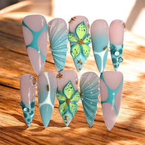 May include: A collection of ten long, almond-shaped artificial nails. The nails showcase diverse designs, including teal and white patterns, sculpted fan-like shapes, and floral embellishments with gold accents. Some nails have small, clear, bubble-like decorations.