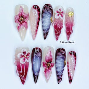 May include: A set of ten long, pointed artificial nails. Each nail features a unique design with a base color of clear or light pink. Designs include floral accents, gradients, and decorative elements in shades of pink, white, and gold.