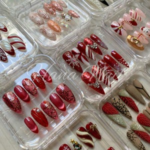 May include: A collection of press-on nail sets in clear plastic storage cases. The almond and stiletto shaped nails feature various holiday designs including red and white candy cane stripes, glittery red finishes, snowflake patterns, and gold accents.