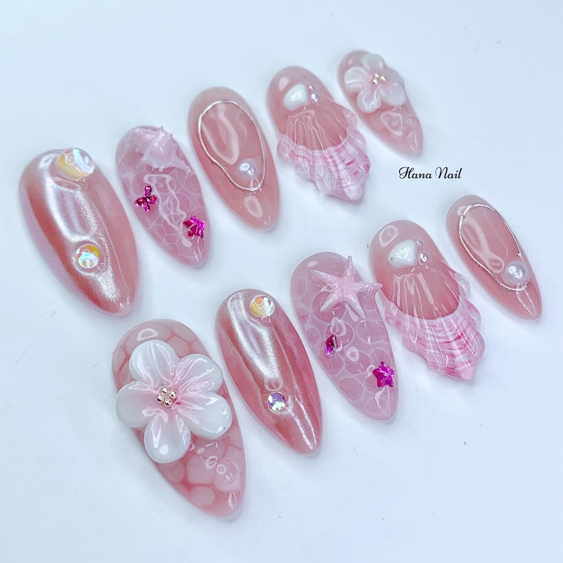 Peut inclure: Ensemble de dix faux ongles en forme d'amande rose et blanc avec diff&eacute;rents motifs sur le th&egrave;me de l'&eacute;t&eacute;, notamment des coquillages, des &eacute;toiles de mer et des fleurs. Les ongles sont d&eacute;cor&eacute;s de paillettes iris&eacute;es et de strass roses. Le texte "Hana Nail" est visible sur l'image.