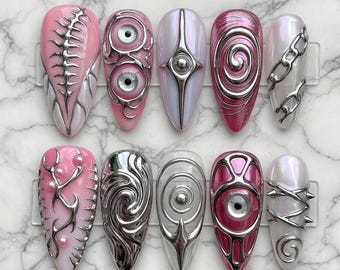 May include: A set of ten long, almond-shaped artificial nails with intricate designs. The nails feature a variety of colors, including pink, silver, and white, with metallic embellishments and unique patterns. The designs include spirals, chains, and eye-like shapes.