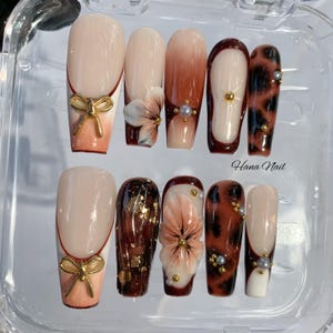 May include: A set of ten artificial nails in various designs. Some nails feature a nude base with gold bow accents, while others showcase floral and leopard print patterns. The text "Hana Nail" is visible on the image.