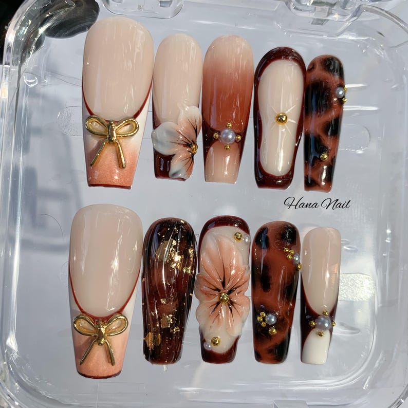 May include: A set of ten artificial nails in various designs. Some nails feature a nude base with gold bow accents, while others showcase floral and leopard print patterns. The text "Hana Nail" is visible on the image.